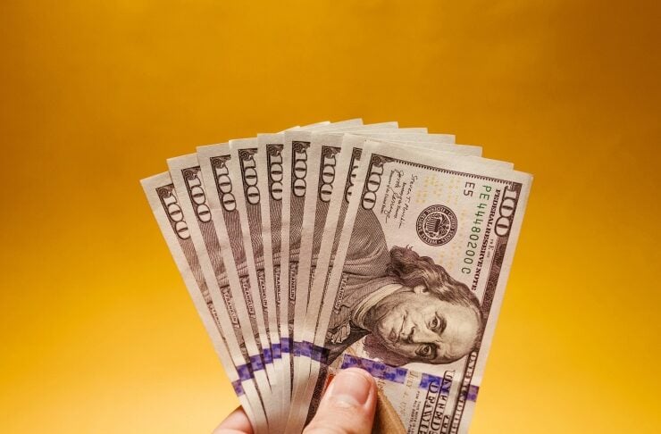 A hand fanning multiple US $100 bills against a bright yellow background.
