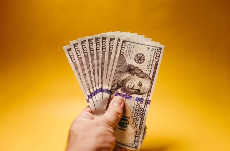 Close-up of a hand fanning multiple $100 bills against a warm yellow background, symbolizing cash.