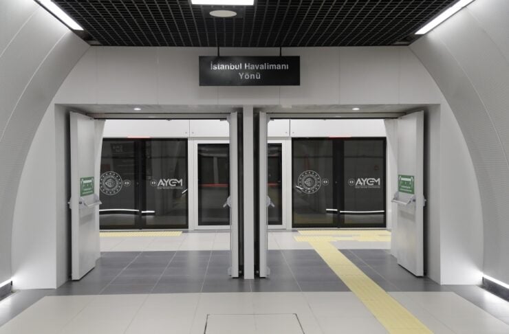 Symmetrical airport rail entrance with two glass doors, overhead sign reading 'İstanbul Havalimanı Yönü' and a yellow floor guiding path.