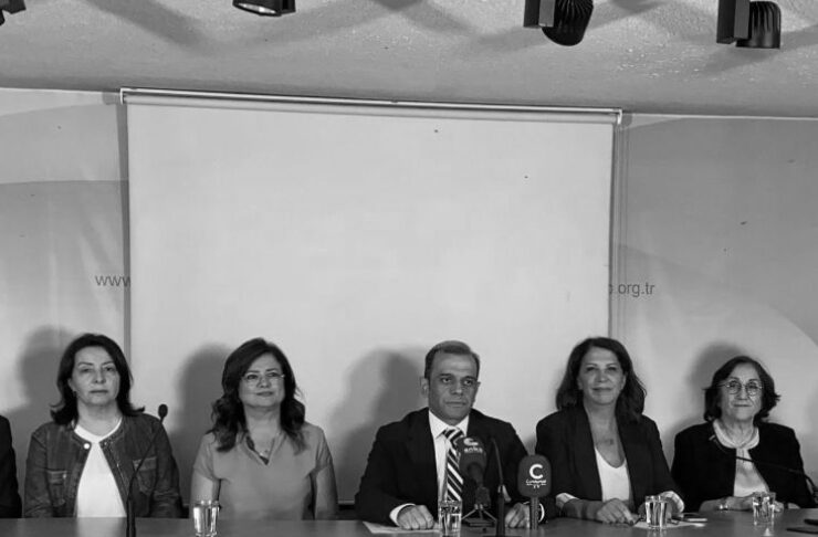 Panel of five people sit behind a curved press desk with microphones and water glasses, in a conference room with a projector screen behind them (black-and-white photo).