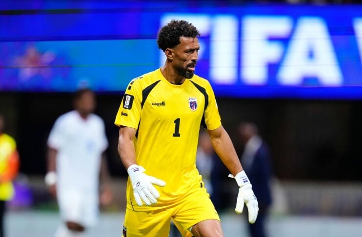 Soccer goalkeeper in a bright yellow kit with number 1, standing on the field with a ball at his feet during a match.