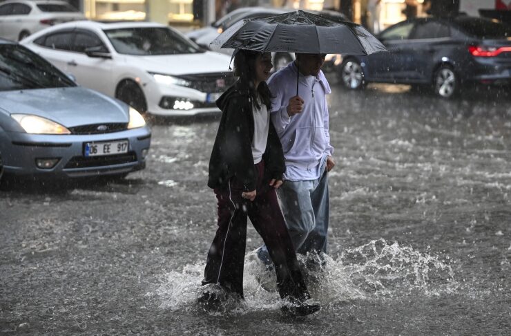Two people share a large umbrella while wading through a flooded city street as cars drive by in heavy rain.