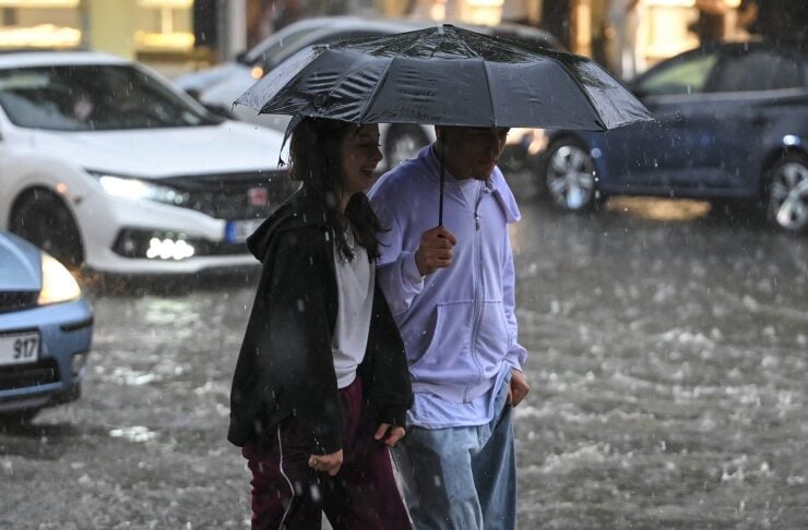 Two people walk through a rain-filled street under a large umbrella as water splashes around their boots.