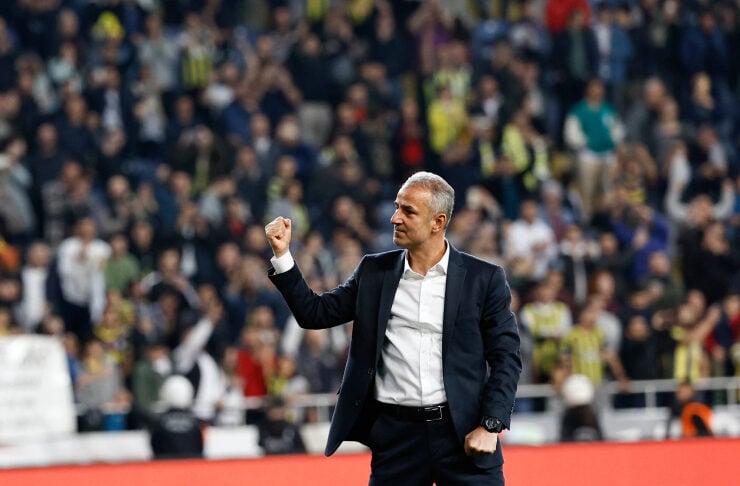 Football coach in a dark suit on the field, fist raised in celebration as fans cheer in the stands behind him.