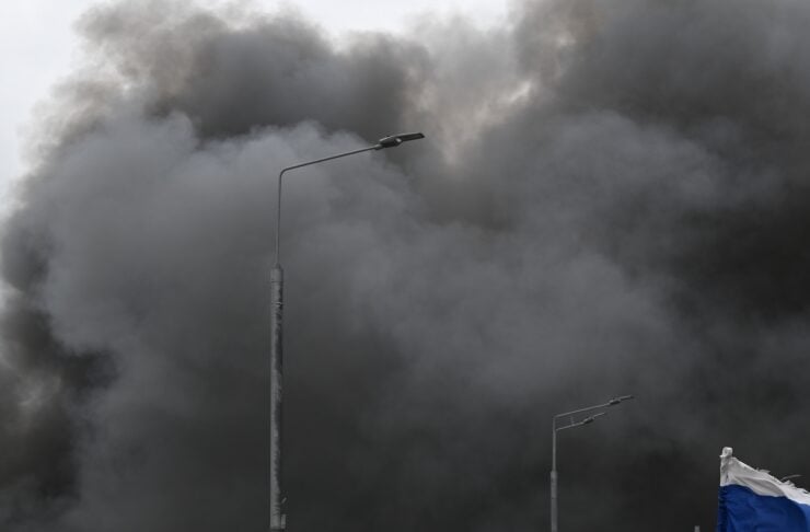 Group of men walking near a car with a Russian flag as a massive smoky cloud rises behind them.