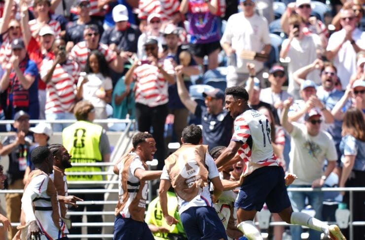 Soccer players in white uniforms celebrate a goal on the field as fans cheer in the stands behind them.