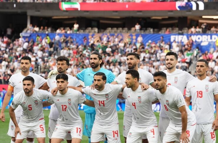 Iran national football team in white kits pose for a group photo on the field, goalkeeper in blue, during a FIFA World Cup event.