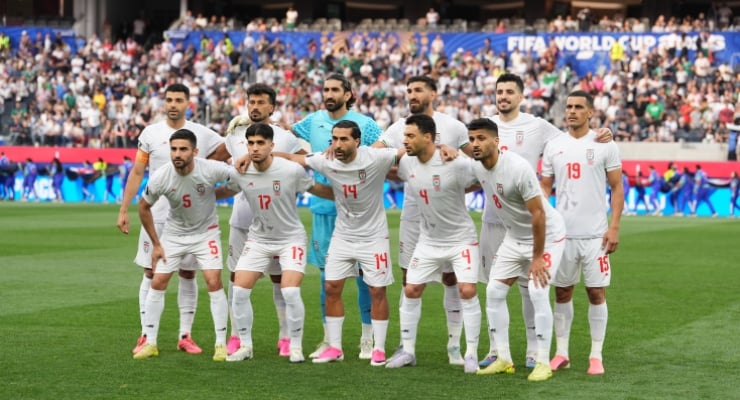 Eleven football players in white kits pose for a group photo on a green pitch before a match, with a crowded stadium in the background.