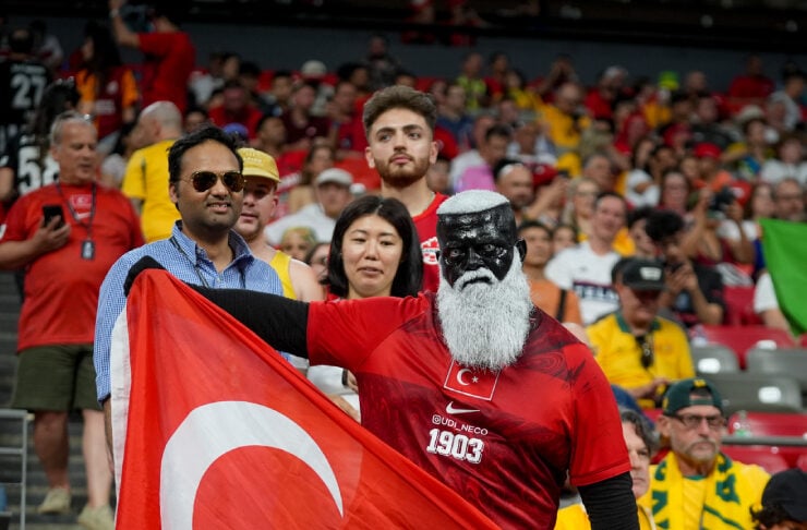 Fan with a black beard mask and red Turkey jersey holding a large Turkish flag in a crowded stadium.
