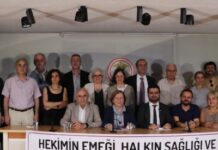 Group of 14 people, seated and standing behind a banner with Turkish text about strengthening TTB for doctors' work and public health, at a panel or press event.