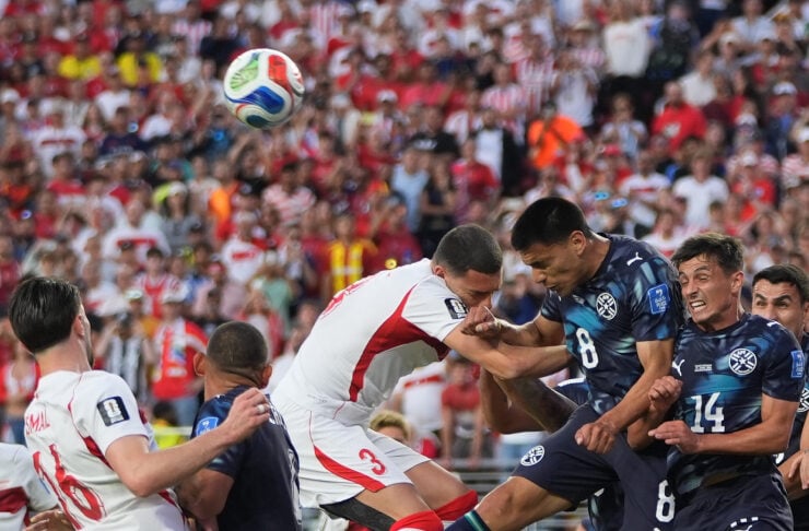 Soccer players from white and dark-blue uniforms jump for a header as the ball flies above the crowd.