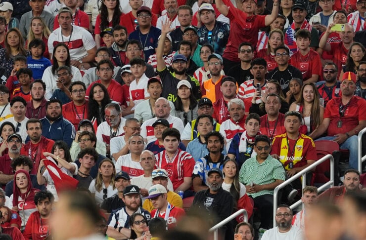 Crowd of soccer fans in red and white jerseys watching a match, with players blurred in the foreground.