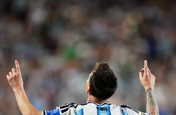 Soccer player Lionel Messi raises both arms with index fingers pointing upward, back facing camera in a striped blue-and-white jersey.