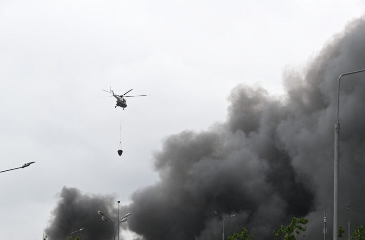 A helicopter hovers over a highway, lowering a bucket toward the ground as a large black smoke plume rises from a nearby fire.