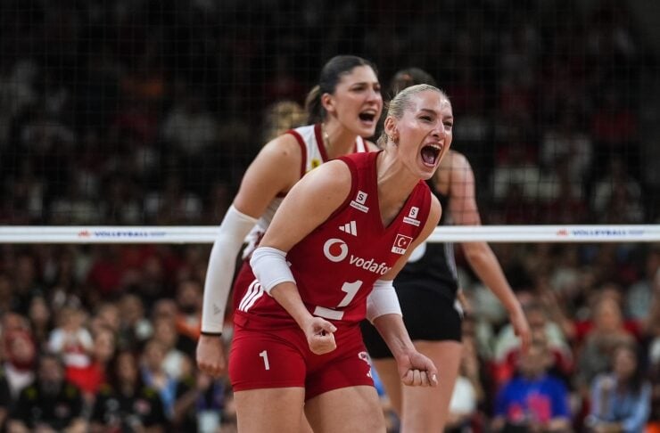 Two female volleyball players in red uniforms celebrate on the court during a match, one leaning forward with a clenched fist in excitement.