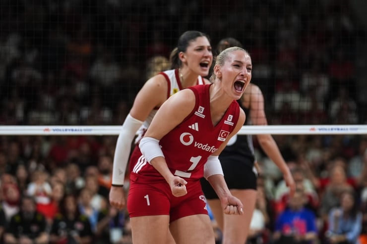 Two female volleyball players in red uniforms celebrate on the court during a match, one leaning forward with a clenched fist in excitement.
