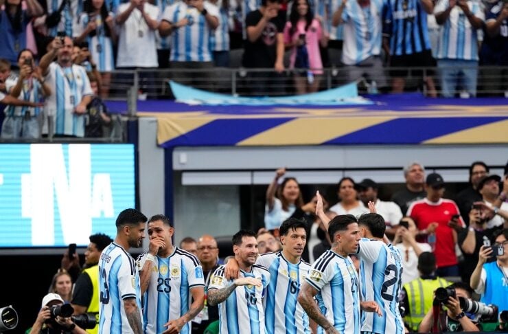 Argentina national soccer players in blue-and-white striped jerseys celebrate on the field as fans cheer in the stands behind them.