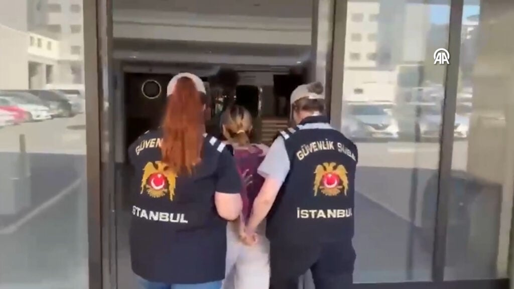 Three security officers in Istanbul vests escorting a person into a glass entrance, back view, urban backdrop behind them.