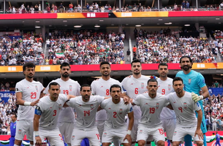 Iran national football team players pose for a group photo on the pitch before a match, wearing white kits with red numbers, with a packed stadium in the background.