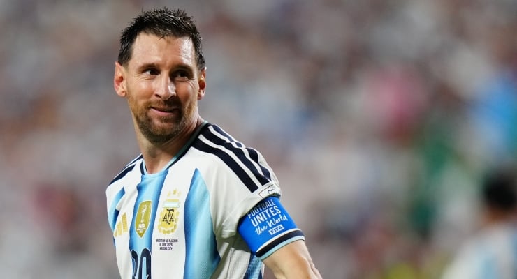 Soccer player in blue-and-white Argentina jersey looking over his shoulder on the field, with a blurred crowd in the background.