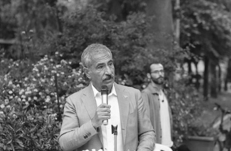 Man in a light suit speaks into a microphone at a transparent podium with flowers, outdoors, at a CHP event.