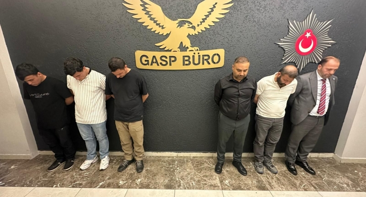 Six men stand with heads bowed against a wall in a police station corridor, beneath a wall sign reading 'GASP BÜRO' and a Turkish police emblem.