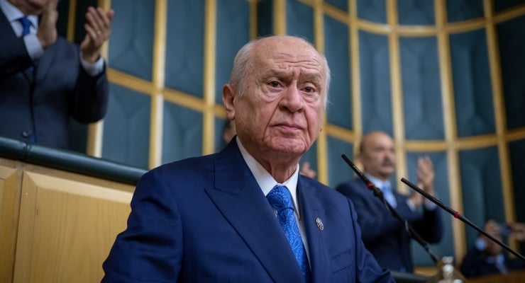 Elderly man in a navy suit and blue tie standing at a podium during a formal assembly event.