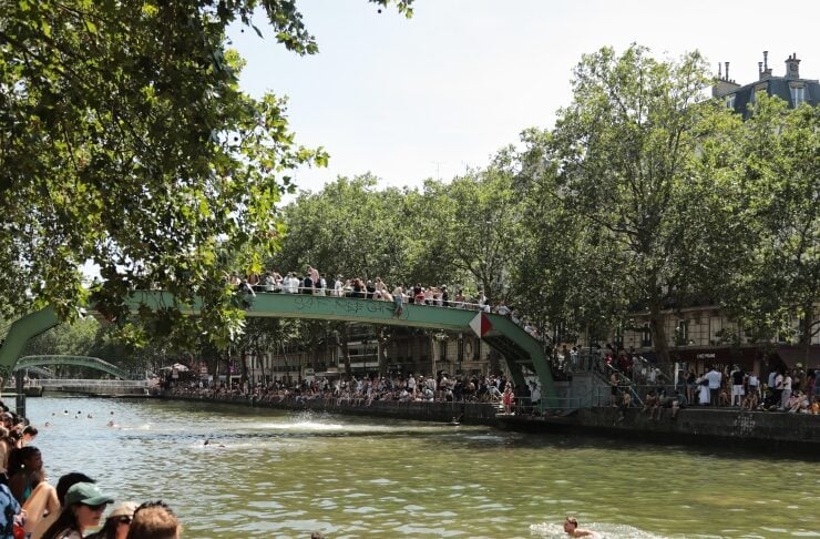 Crowded riverside with people sitting on a green arch bridge and swimmers in a muddy river beneath, shaded by trees on a sunny day close to the bank.