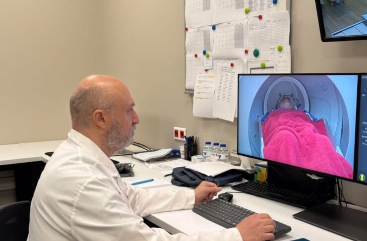 Male clinician in a white lab coat sits at a desk, reviewing an MRI scan on a dual-monitor workstation with a patient in the MRI bore visible on the screen.