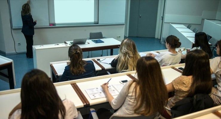 Instructor standing at the front teaching a lecture, while students sit in tiered desks taking notes in a university classroom with a projector screen behind.