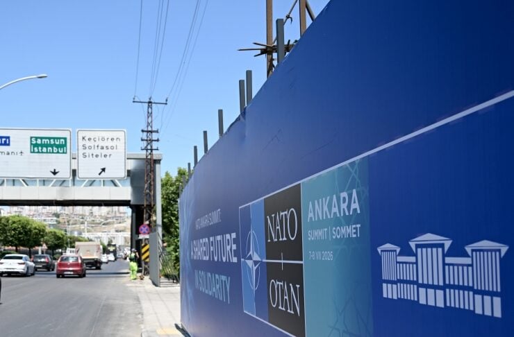 Blue construction barrier wall with NATO Ankara Summit signage along a sidewalk; city street with cars and overhead road signs in the background