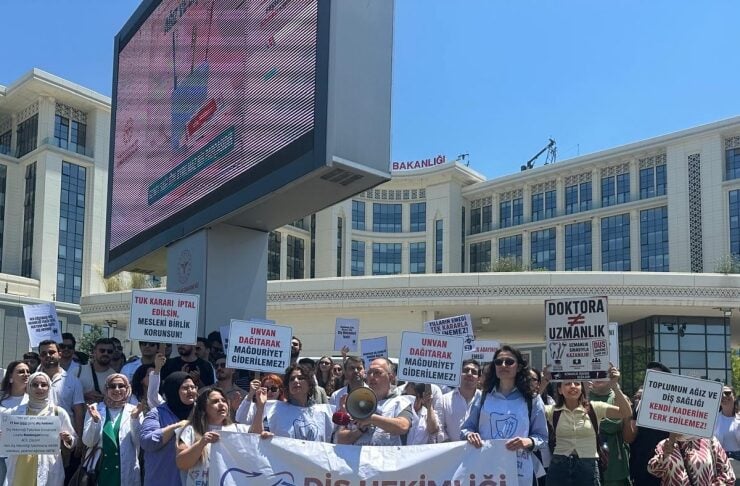 Group of healthcare workers protesting outdoors, holding a large banner that reads 'Diş Hekimliği Sendikası' while others hold signs in Turkish, in front of a modern government-style building with a large digital screen.