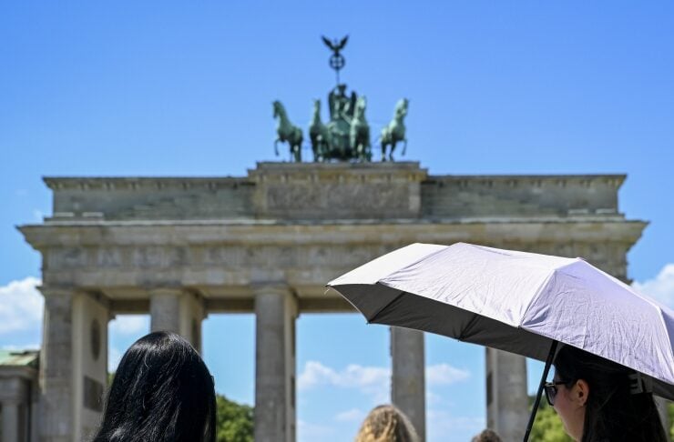 Two women walk toward the Brandenburg Gate on a sunny day; one with long black hair reaches toward a friend holding a lavender umbrella.