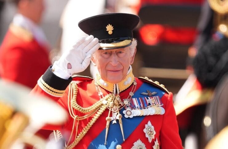 Older man in ceremonial red uniform saluting during a parade, wearing medals and a peaked cap.