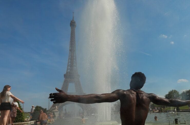 Man standing in a shallow fountain with arms outstretched as water jets shoot up; a tall Eiffel Tower-like structure visible in the background on a sunny day, with people by the pool edge nearby.
