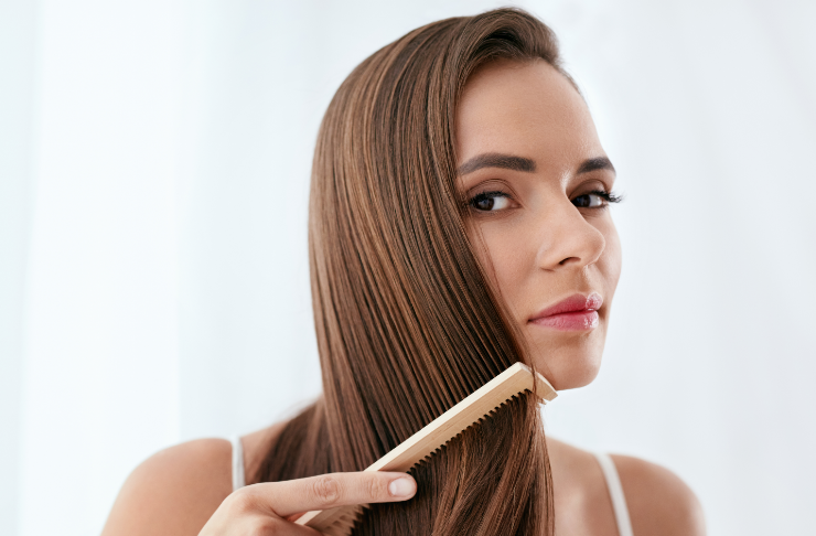 Woman with long brown hair brushing it with a wooden comb, looking at the camera.