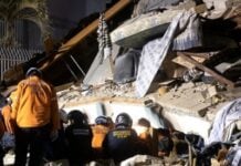 Rescue workers in orange jackets and helmets search through a collapsed building with rubble and hanging fabric pieces on top of debris.