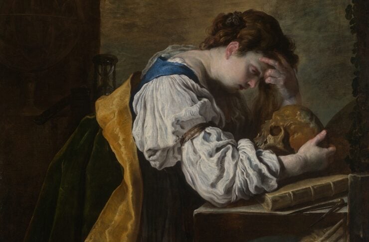 A woman in a white blouse leans on a desk, resting her head on her hand beside a skull, in a somber vanitas scene.
