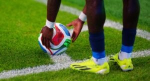 Two soccer players place a multicolored ball on the grass near a sideline line, preparing for a restart or kick-off, with bright cleats visible.