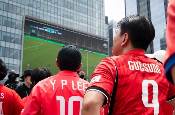 Crowd of fans in red jerseys looking toward a large stadium screen during a soccer match, with players visible on the field in the distance. The jersey on the left reads 'Y P LEE' and number 10, while the one on the right reads 'GUESUNG' and number 9.
