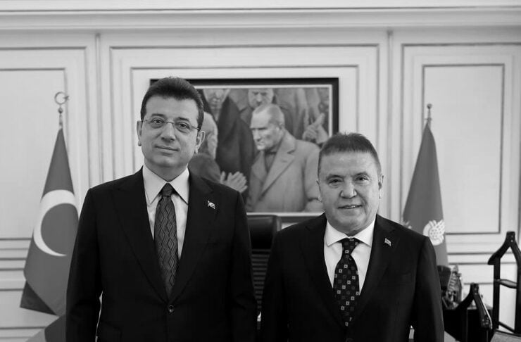 Two men in suits stand together in a formal office with Turkish flags visible behind them.