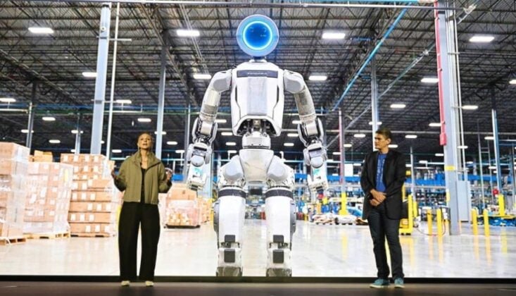 Tall white humanoid robot with a glowing blue head stands between two people in a spacious warehouse demo area at a factory.