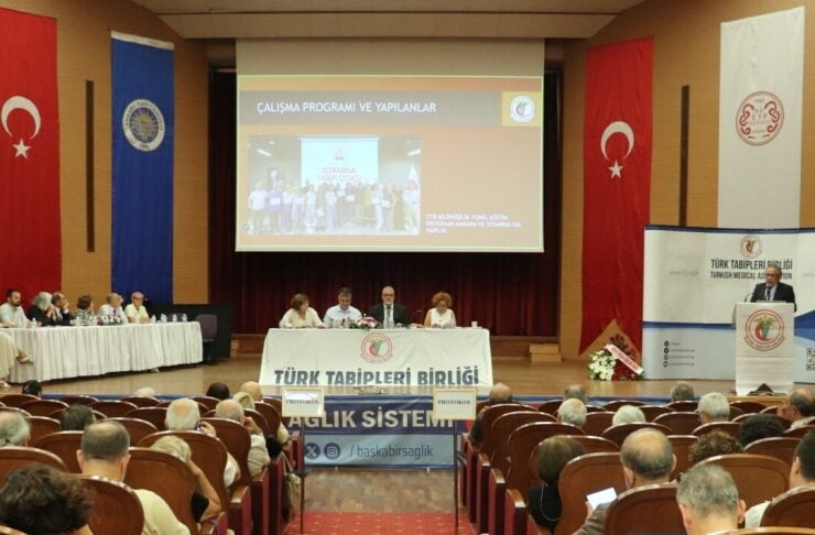 Conference hall conference with a panel at a long table on stage, Turkish Medical Association banners, and flags flanking the screen.