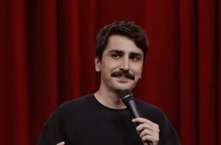 Male comedian with a dark mustache in a black sweatshirt, speaking into a handheld microphone on a stage with red curtains behind him, mid-performance.
