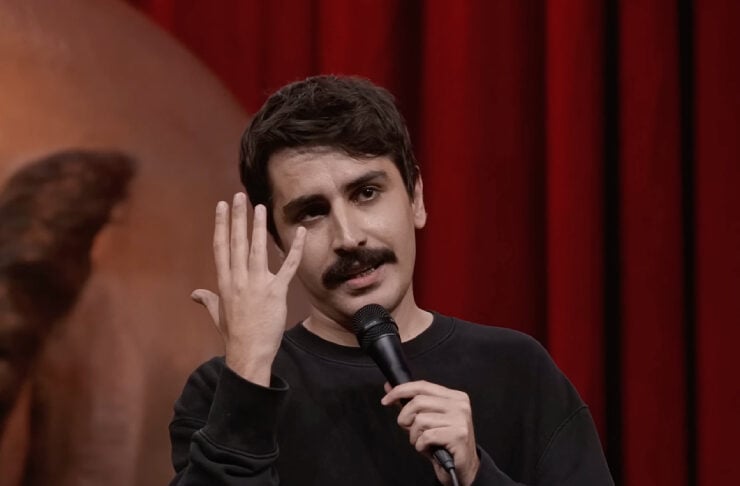 Comedian on a stage with a microphone, gesturing with his raised hand against a red curtain backdrop.