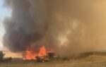 Rural field burning with large orange flames and thick black smoke; a yellow firefighting truck is nearby fighting the fire.