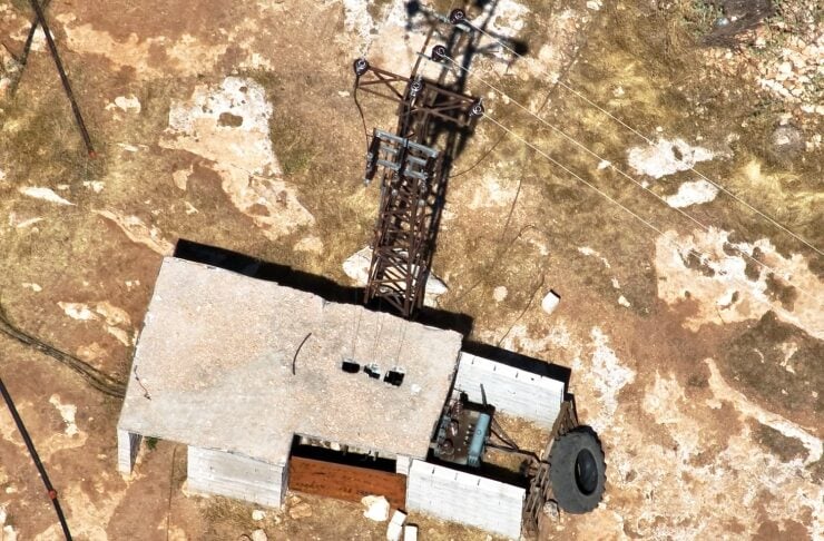 Aerial view of a small rectangular building with a tall lattice tower and guy wires on dry, rocky terrain; a large tire sits near the side of the structure.