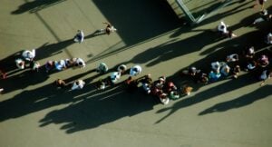 Aerial view of a long line of people standing on a concrete surface, casting long shadows in the sun.