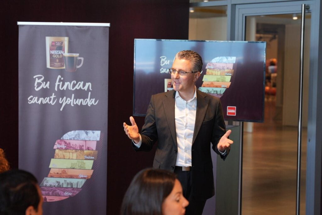 Man in a suit giving a presentation beside a banner and a screen in a conference setting