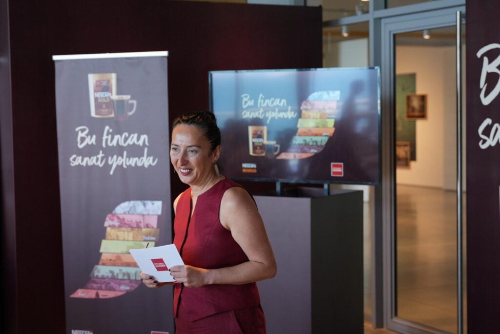 A woman in a burgundy sleeveless dress speaks to an audience while holding papers at a promotional booth with banners and a screen behind her.